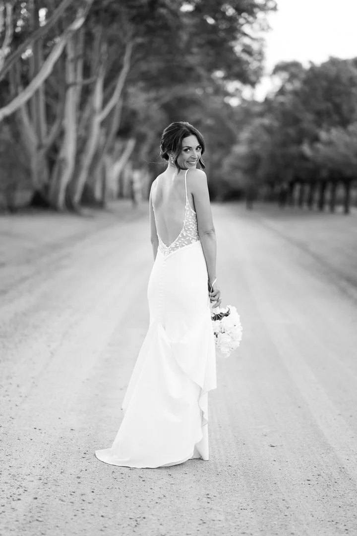 A woman in a wedding dress standing on a tree-lined dirt road, holding a bouquet, smiling at the camera.