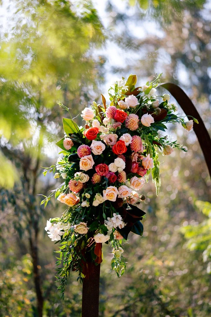A floral arrangement with pink, white, and red roses, dahlias, and greenery hanging on a black metal stand outdoors.