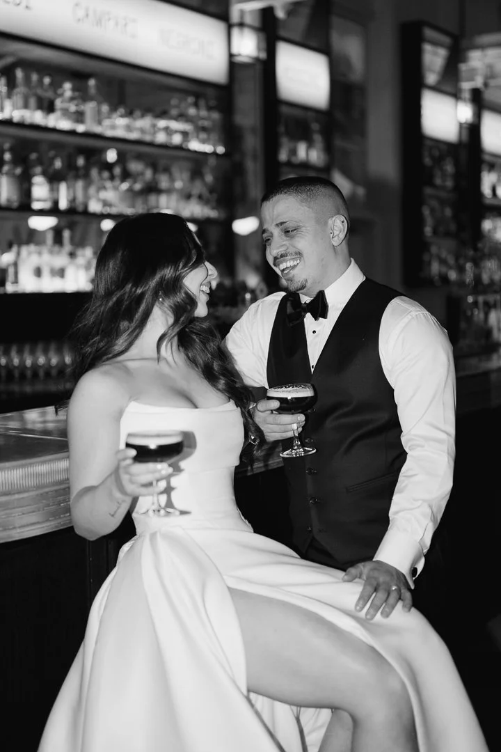A woman in a white strapless dress and a man in a tuxedo with a vest and bow tie sitting at a bar, holding drinks and smiling at each other.