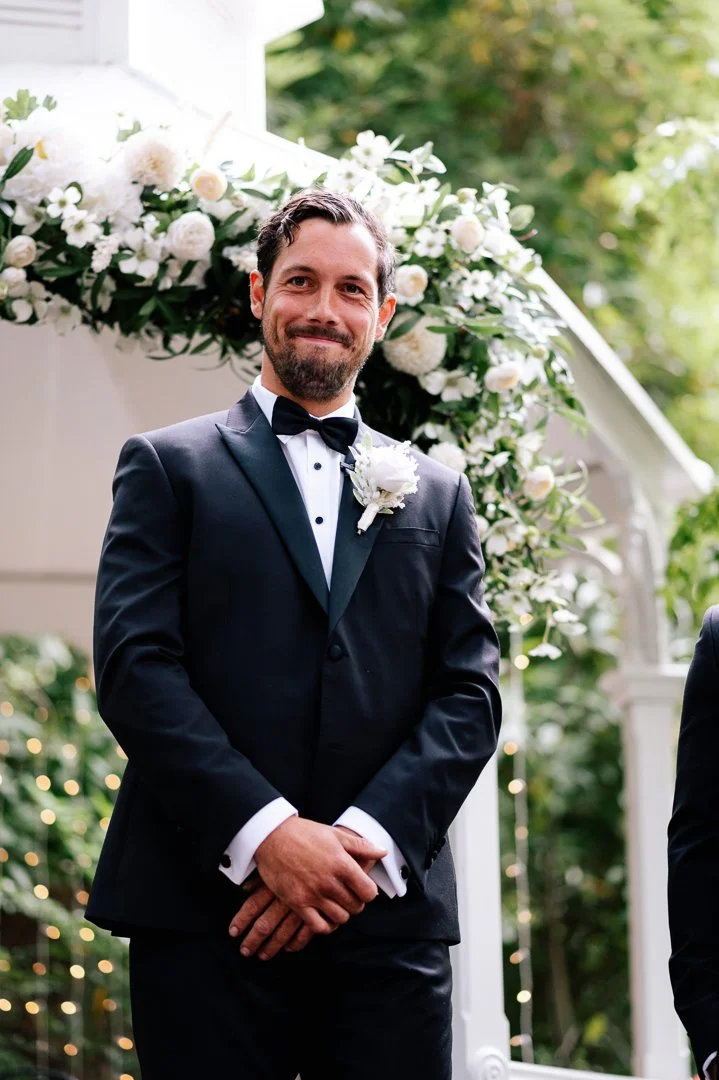A man in a black tuxedo, white dress shirt, and black bow tie, standing outdoors during a wedding ceremony, smiling with hands clasped in front of him, with a floral arch decorated with white roses and greenery in the background.