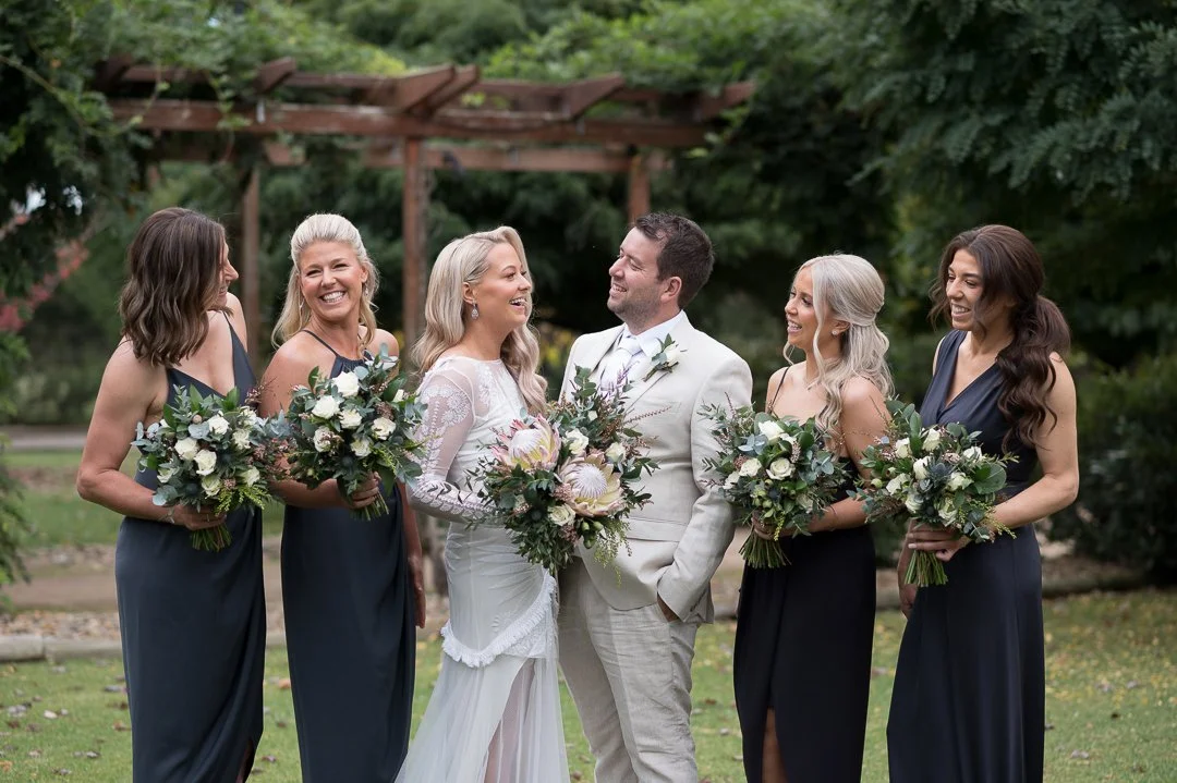 A wedding party outdoors with a bride, groom, and four bridesmaids holding bouquets of flowers, standing on grass with greenery and trees in the background.