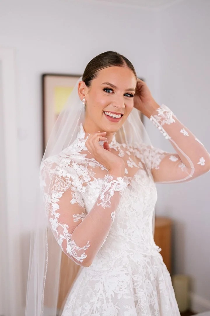 A bride in a lace wedding dress smiling and adjusting her hair in a room.