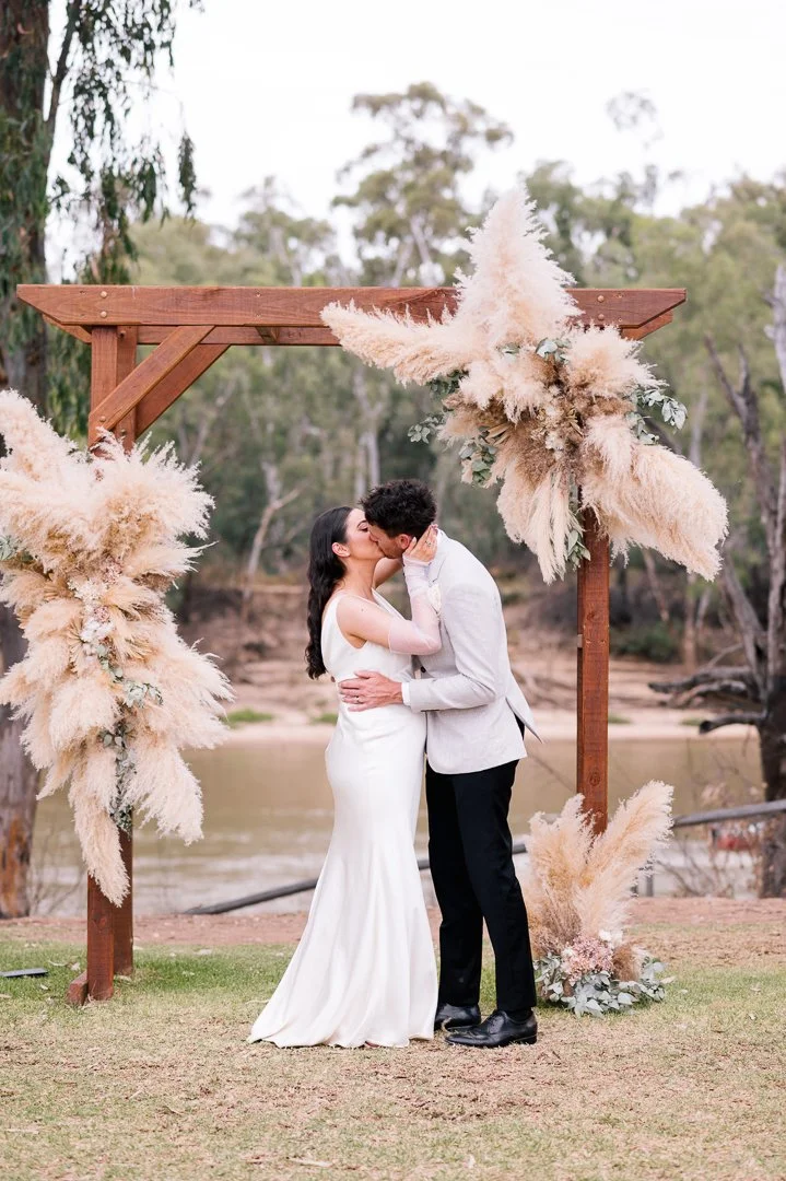 A bride and groom kissing during their outdoor wedding ceremony, standing under a wooden arch decorated with pampas grass and floral arrangements, by a river with trees in the background.