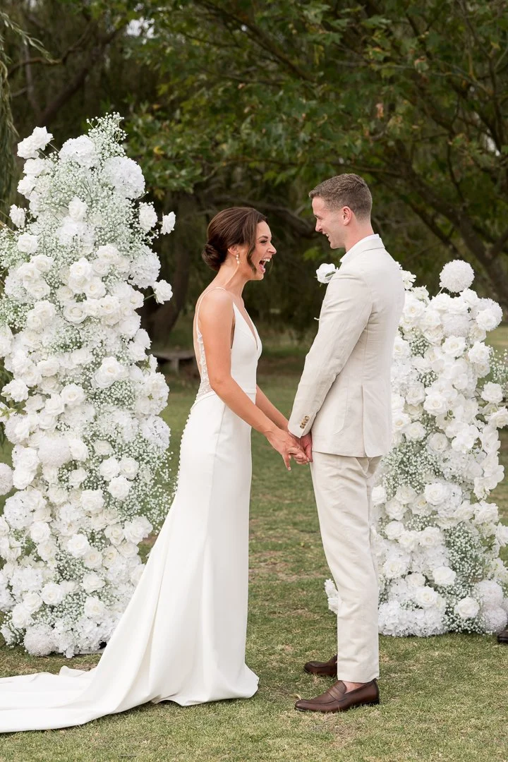 Bride and groom holding hands, smiling, standing outdoors near white floral arch at wedding ceremony.