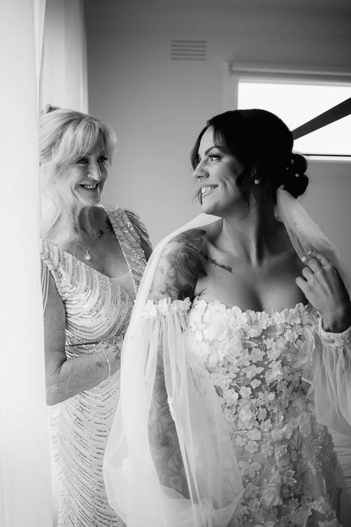 A bride with tattoos and a veil looking at a smiling woman in a beaded dress, both in a room with a window.