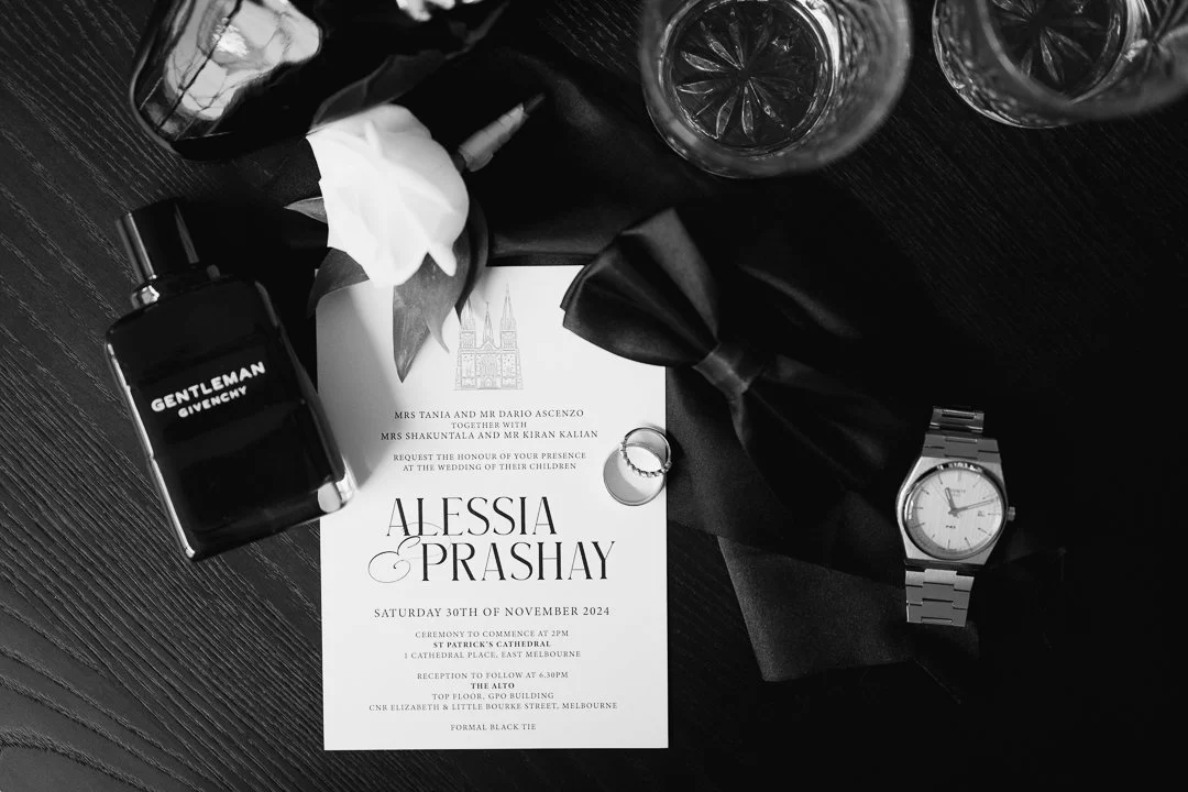 Wedding invitation surrounded by a perfume bottle, white flower, wedding rings, a tuxedo bow tie, a watch, and glasses of water on a black wooden surface.