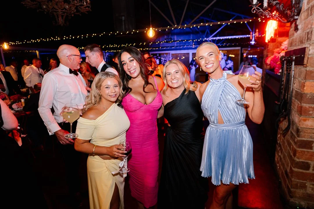 Four women in colorful dresses smiling and holding drinks at a lively celebration or party in a decorated indoor venue.