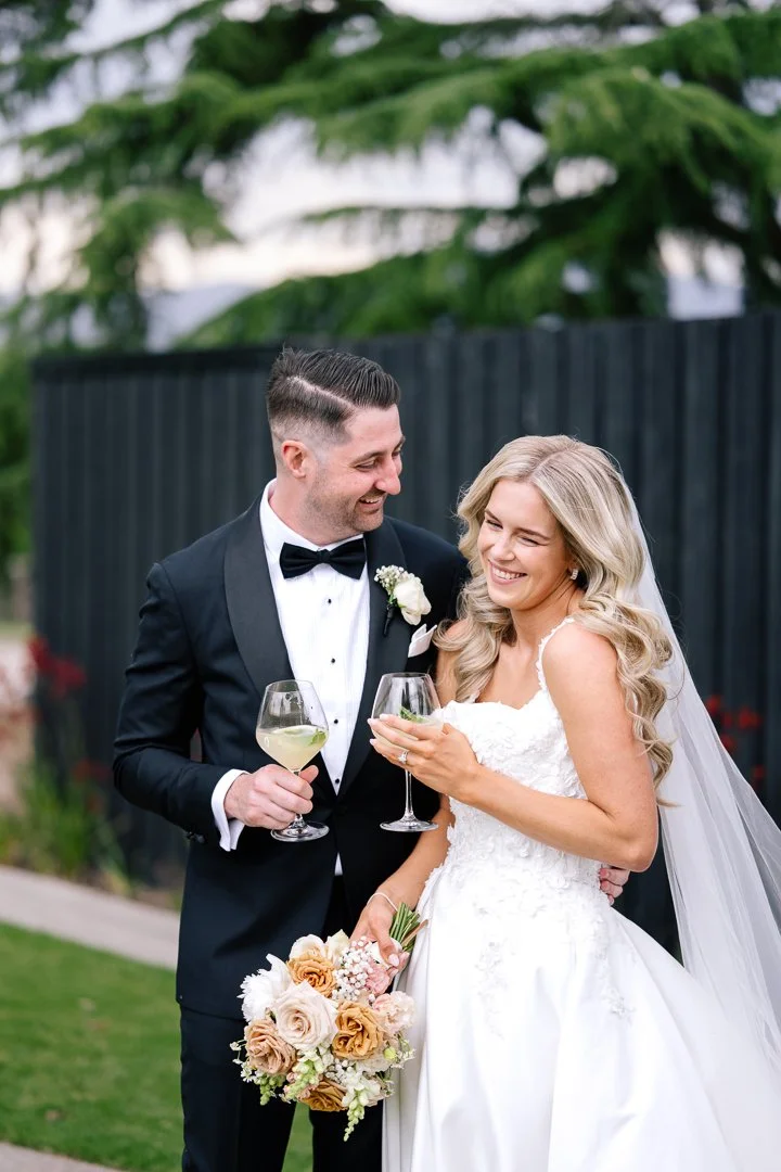 A newlywed couple in wedding attire sharing a toast with wine glasses outdoors, holding a bouquet, smiling at each other, in front of a black fence and green trees.