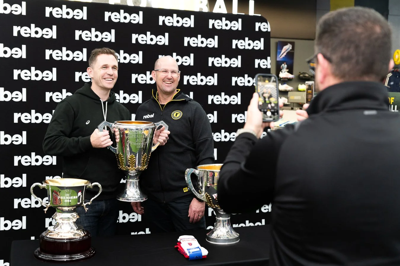 Two men smiling and holding a large trophy in front of a 'rebel' branded backdrop, with a third person taking a photo of them, and additional trophies on a table in the foreground.