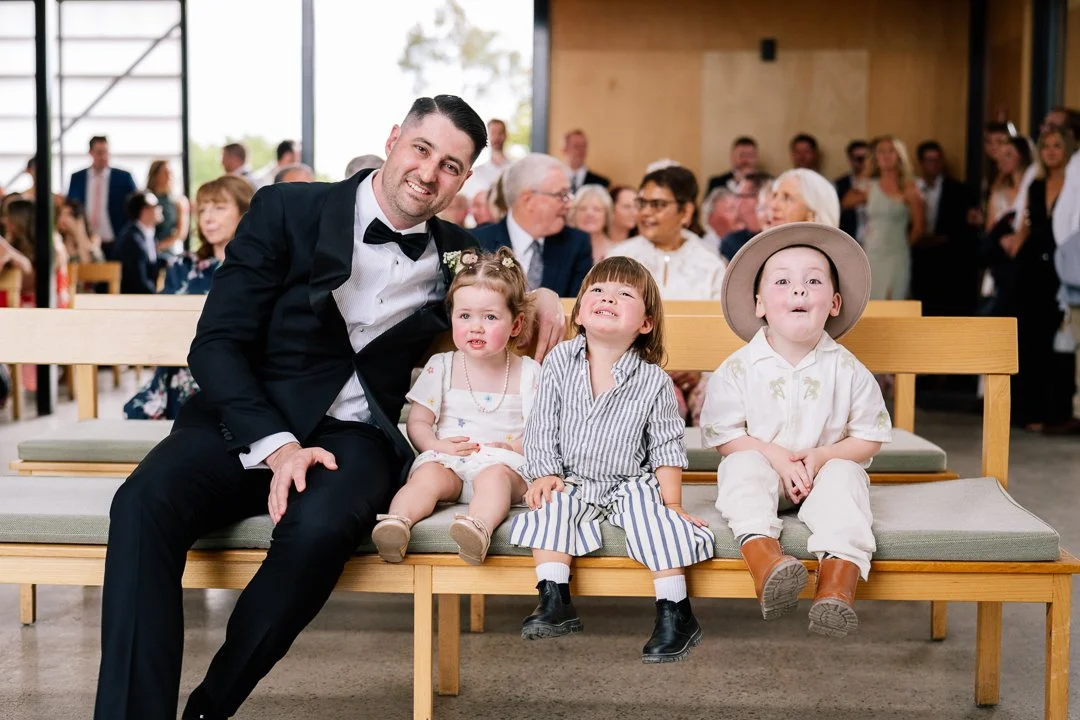 A man in a tuxedo sitting on a bench with three children dressed in vintage clothing, at a formal event with many guests in the background.