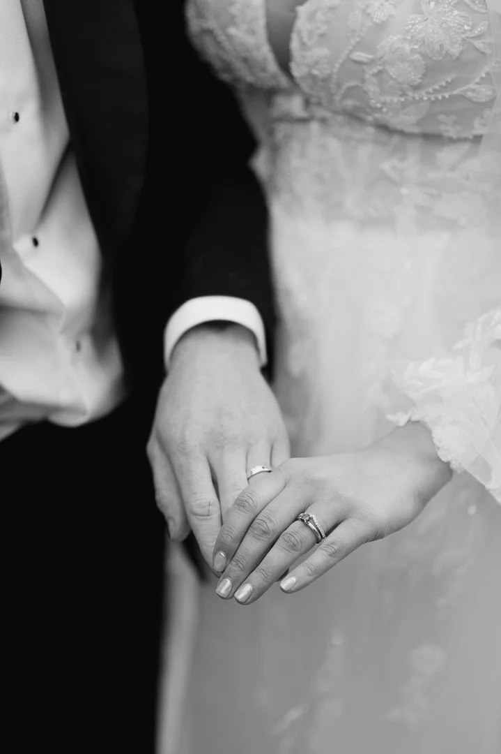 A close-up of a couple holding hands, displaying wedding rings, with the bride wearing a lace wedding dress and the groom in a tuxedo.