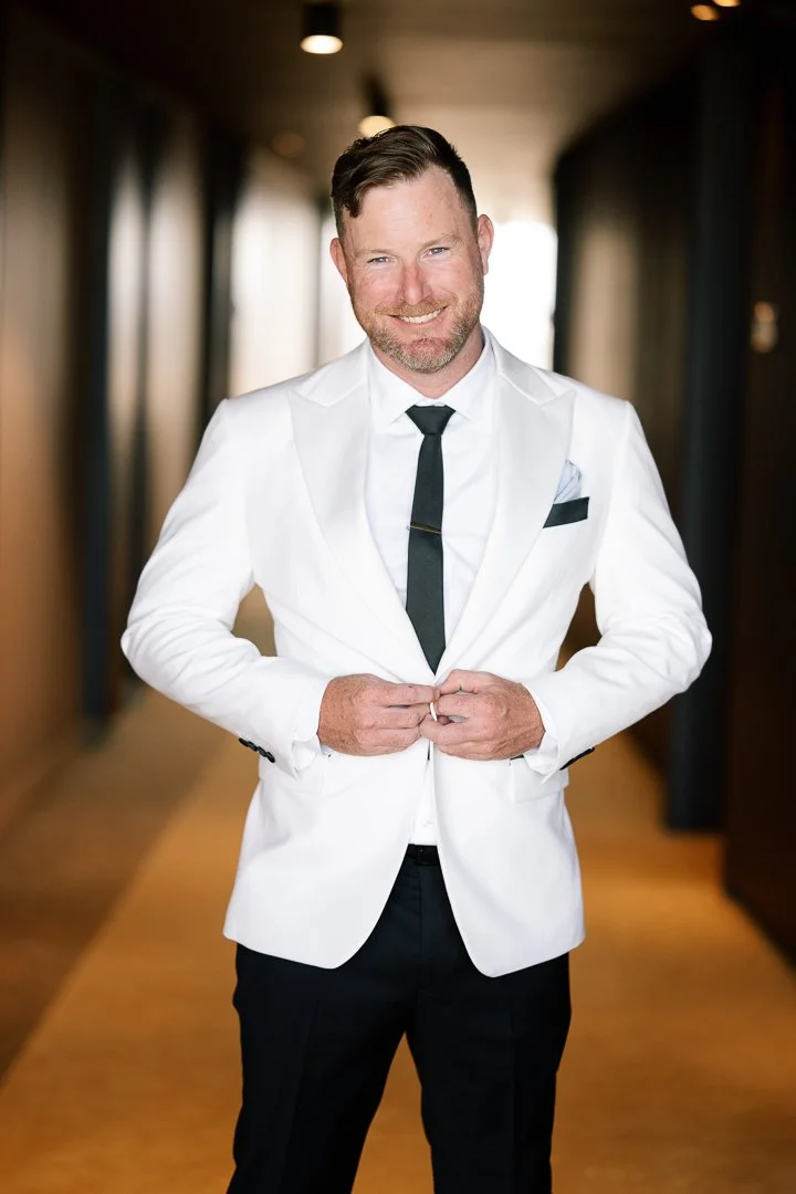 A man in a white tuxedo jacket, white shirt, and black tie stands in a hallway, smiling and adjusting his jacket.