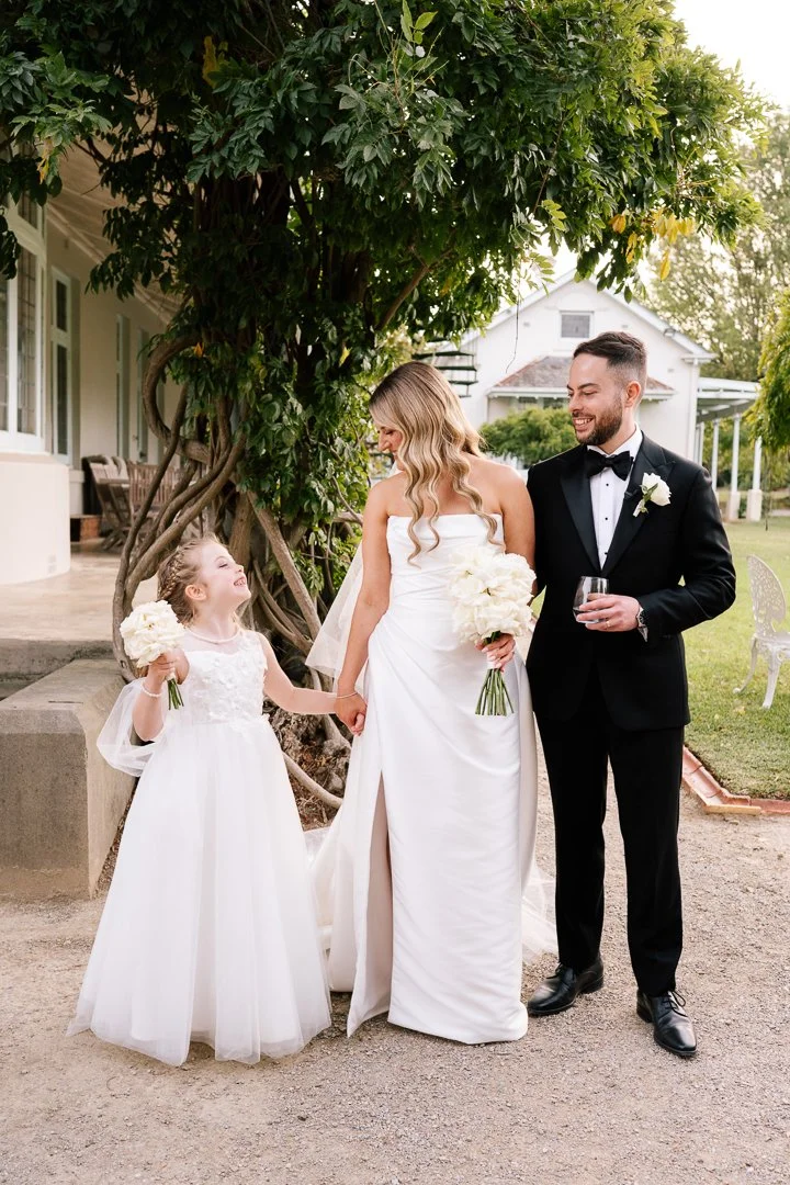 A bride in a white wedding gown, a groom in a black tuxedo, and a young girl in a white dress holding bouquets, standing outdoors near a large tree and a house, smiling and holding hands.