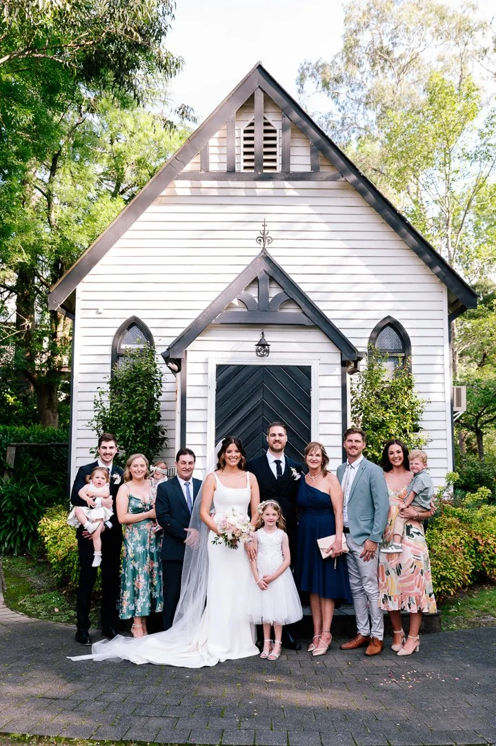 A group of people, including a bride and groom, standing in front of a white chapel, at a wedding celebration.