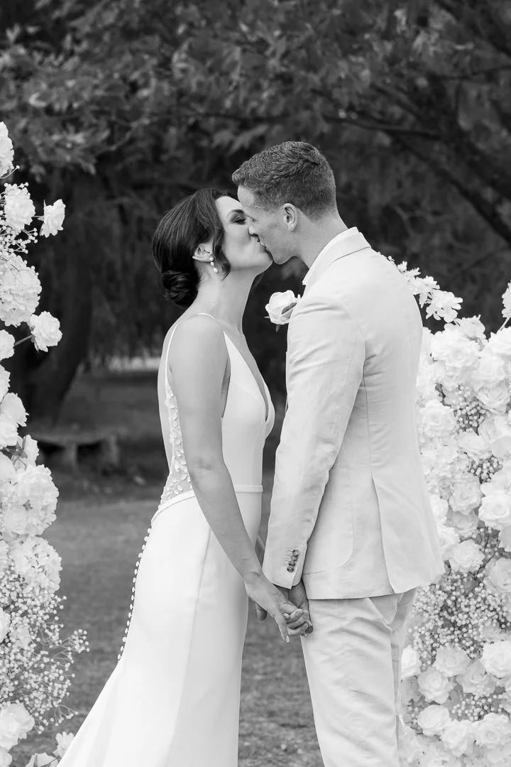 A black-and-white photo of a bride and groom sharing a kiss during their wedding, holding hands, with floral decorations in the background.