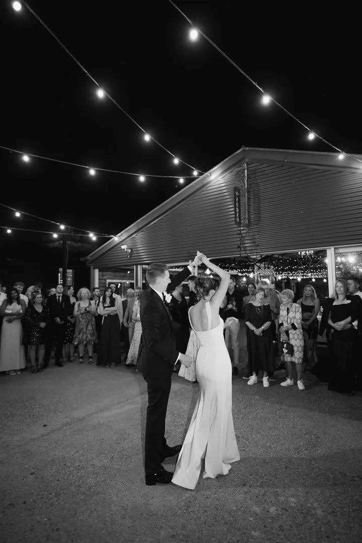 A bride and groom dancing at their wedding reception outside under string lights with guests watching.