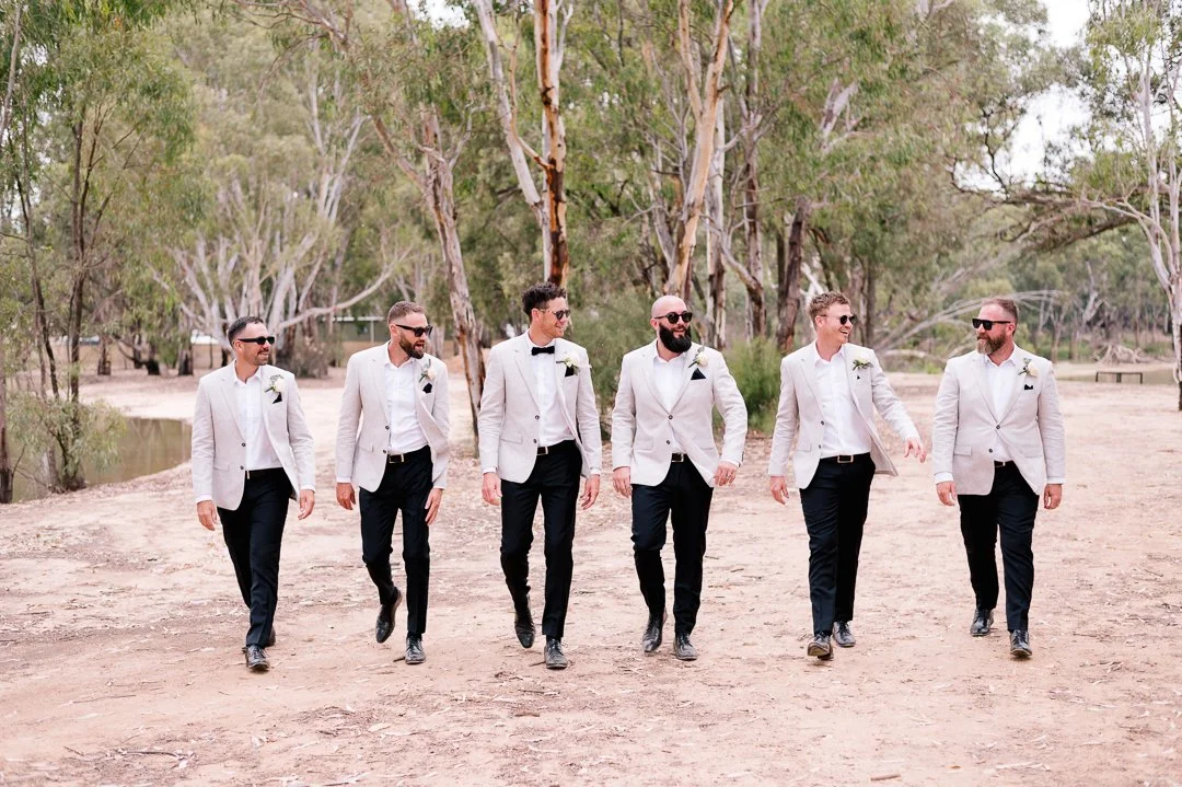Six men dressed in white blazers and black pants walking in a line outdoors with trees in the background.