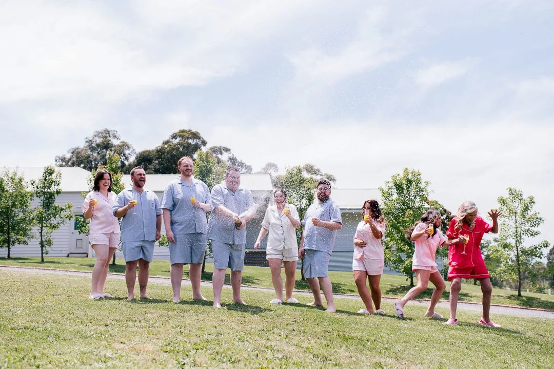 A group of people celebrating outdoors, spraying champagne and holding glasses on a sunny day.