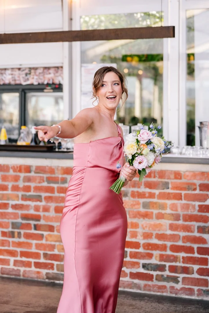 A woman in a pink satin dress holding a bouquet of flowers, pointing and smiling at a social event or wedding reception with a brick wall and bar in the background.