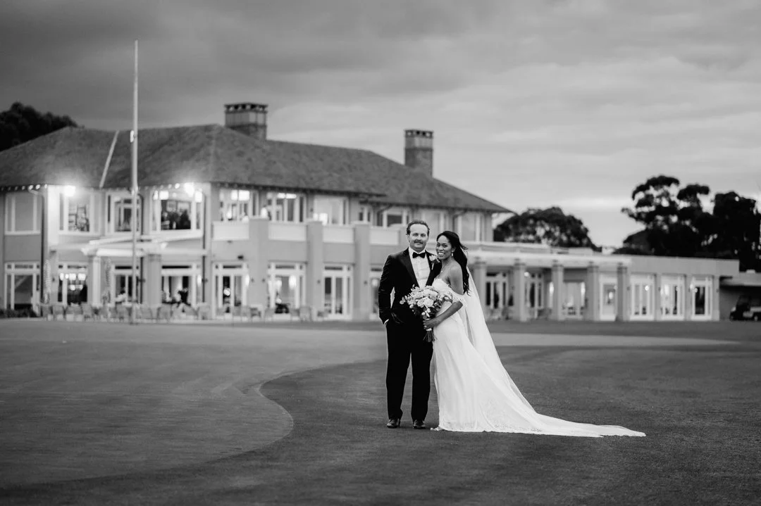 A bride and groom standing together on a golf course in front of a large house, smiling at the camera, with the bride holding a bouquet of flowers.