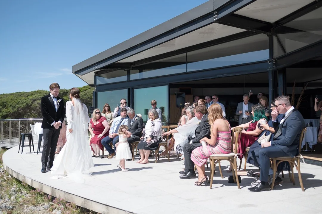 A wedding ceremony taking place outdoors with guests seated on chairs, a bride and groom standing in front of the congregation.