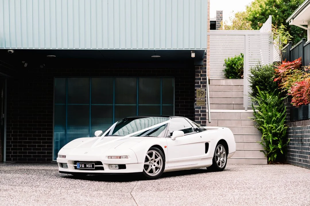 White sports car parked in front of a modern house with stairs and greenery.