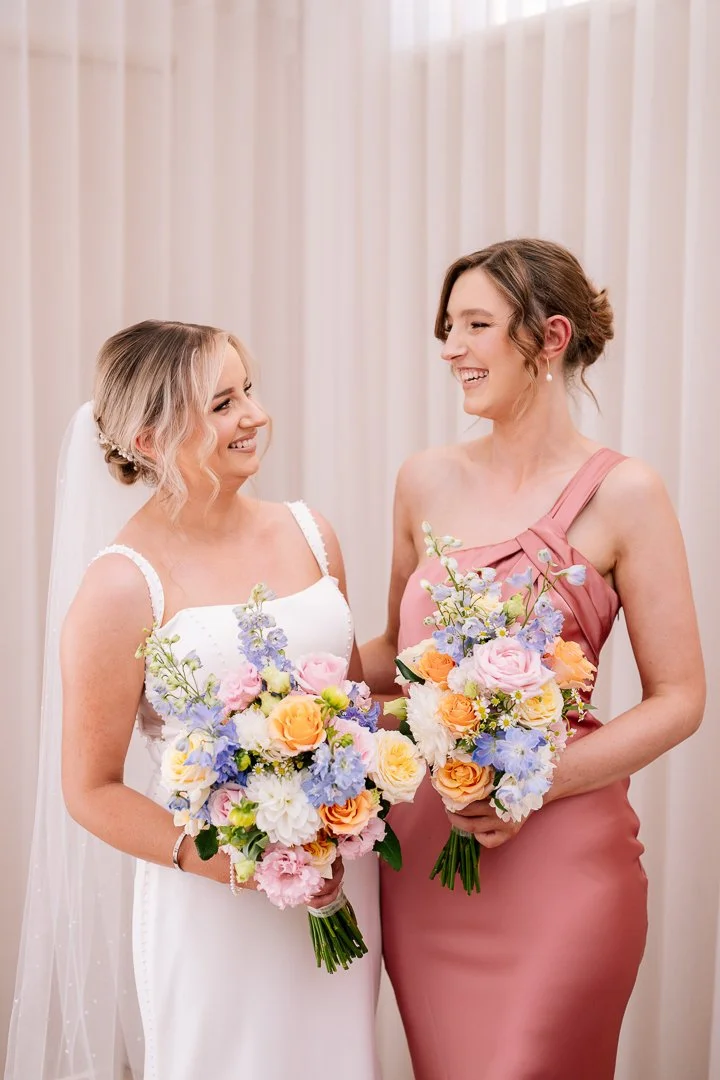 Two women, one in a wedding dress and the other in a pink dress, smiling and holding bouquets at a wedding reception.