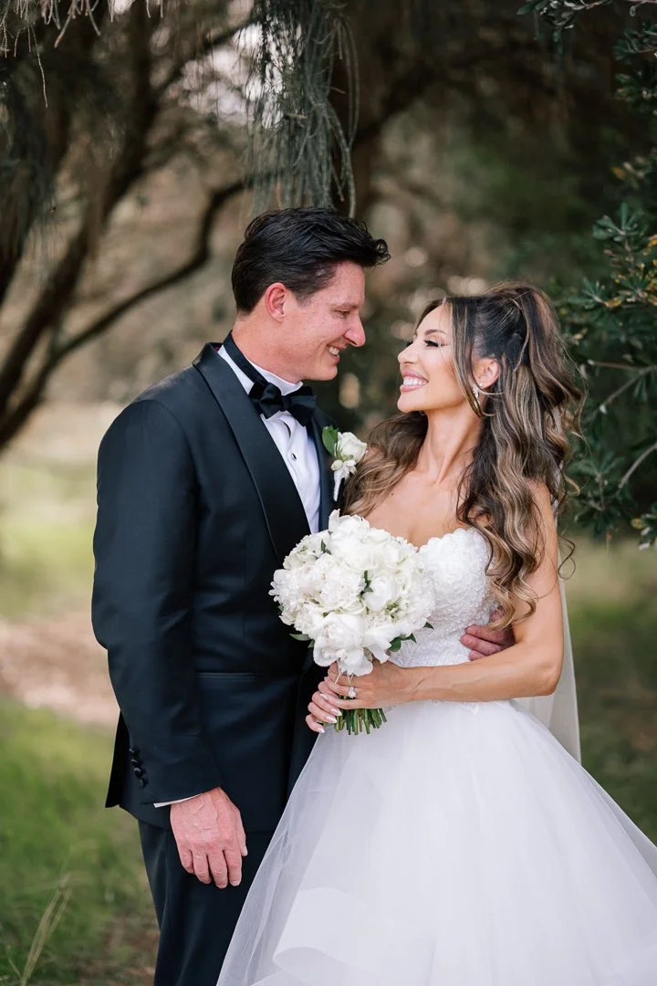 A bride and groom smiling and looking at each other outdoors, with trees in the background. The bride is holding a bouquet of white flowers, and the groom is wearing a black tuxedo with a bow tie.