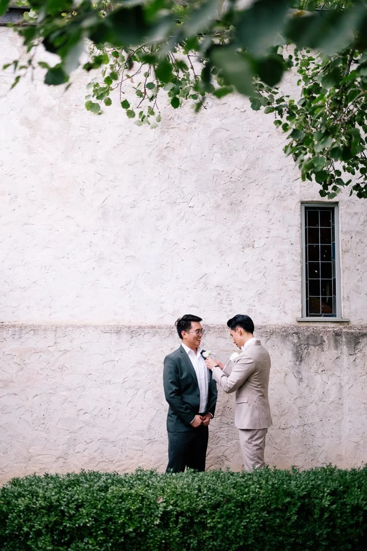 Two men in suits stand outdoors in front of a beige textured wall, with one fixing a boutonniere on the other's lapel, surrounded by green shrubbery and tree branches overhead.
