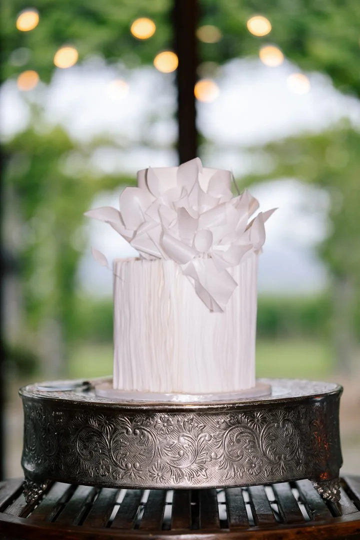 A white cake with textured vertical lines and large white paper flowers on top, placed on an ornate silver cake stand, with blurred green trees and hanging lights in the background.