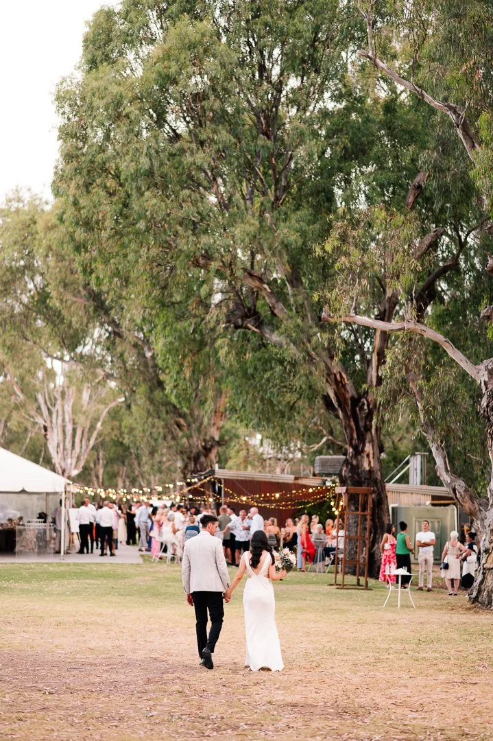 A bride and groom walking hand in hand on a grassy area, surrounded by trees, at their outdoor wedding reception with guests gathered under string lights in the background.