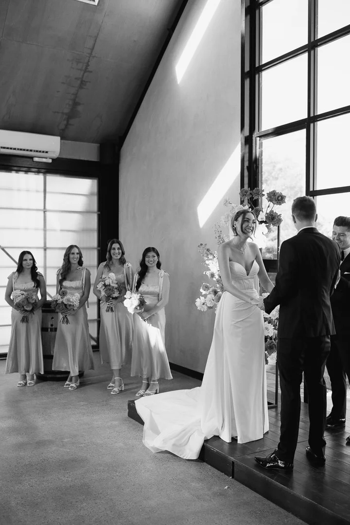 A wedding ceremony with a bride and groom standing on a small platform, holding hands and smiling at each other. The bride is wearing a strapless wedding dress and the groom is in a black suit. Behind them are five bridesmaids holding bouquets, stand