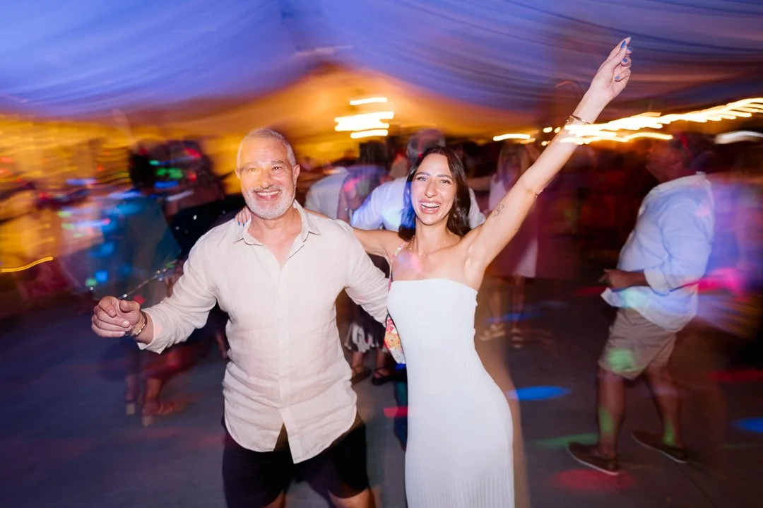 Two people dancing and celebrating at a lively party or event with colorful lights and a blurred crowd in the background.