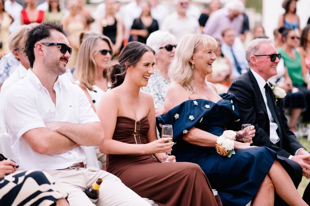 Guests at a wedding or social event, sitting outdoors, dressed in formal attire, some wearing sunglasses, and holding glasses of champagne.