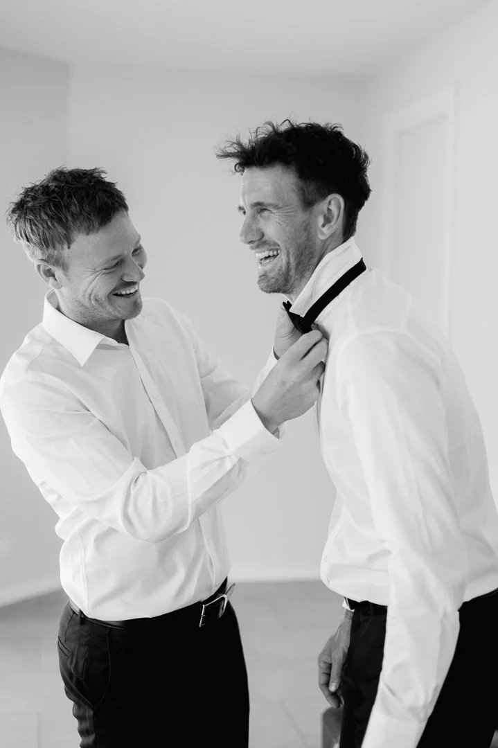 Two men, dressed in white shirts, are smiling and laughing as one helps the other with his bow tie in a bright room.