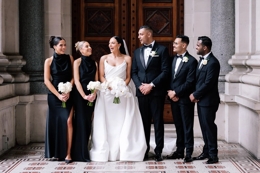 A wedding party of seven people, including a bride in a white wedding dress, a groom in a black tuxedo, three bridesmaids in black dresses, and two groomsmen in black suits, standing together inside a church or cathedral with ornate wooden doors in t