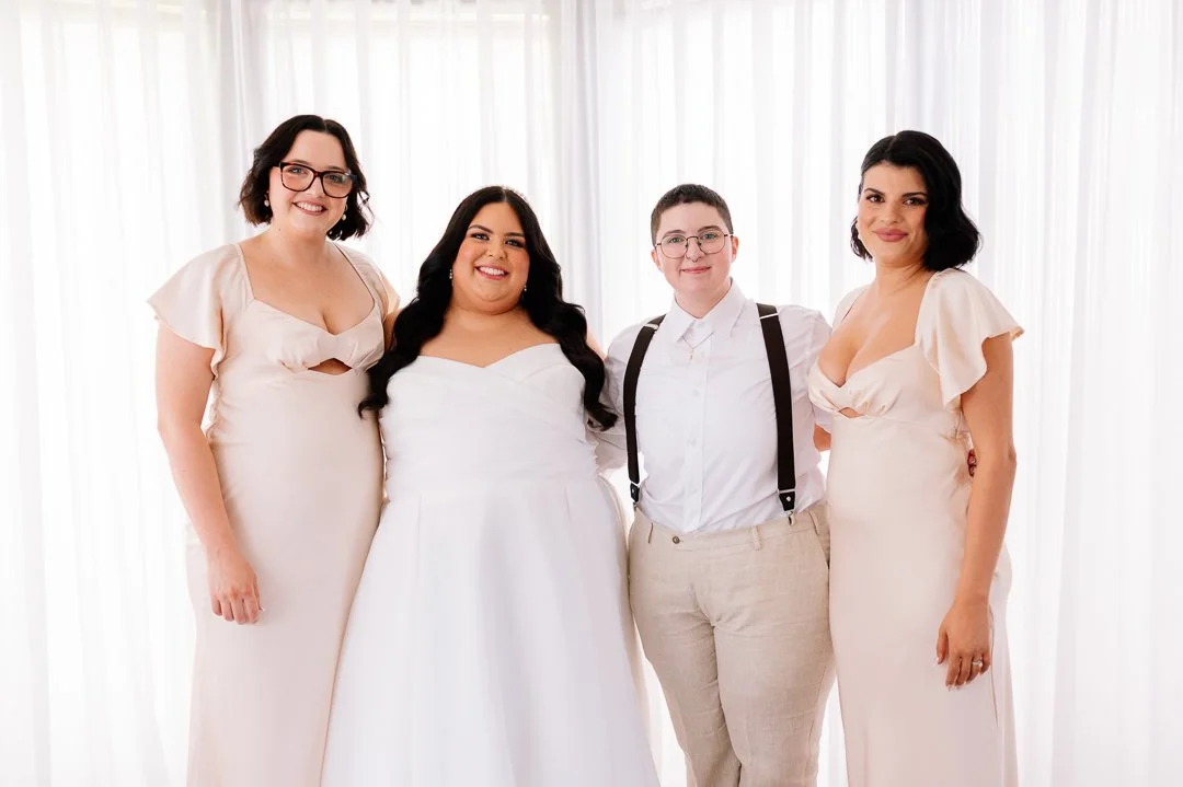 Four women standing together in front of a sheer white curtain, smiling at the camera. One woman is wearing a wedding dress, another in an off-white dress, and the other two in white shirts with beige pants and suspenders.