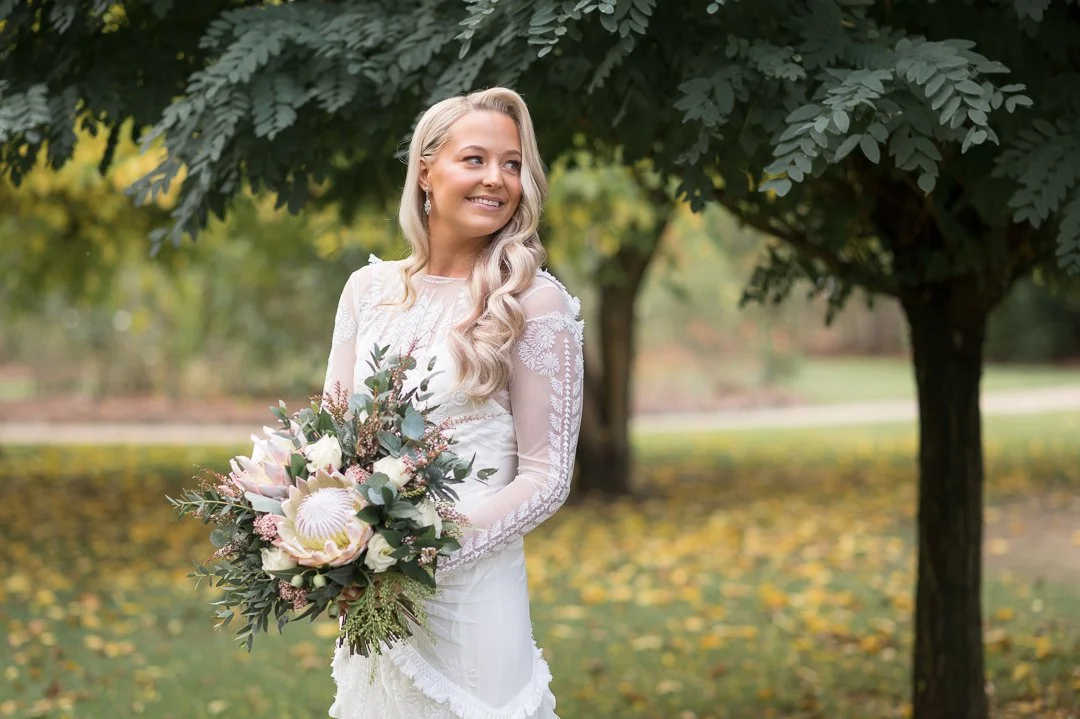 A woman in a white wedding dress holding a bouquet of flowers, standing outdoors near a tree with green leaves and fallen autumn leaves on the ground.