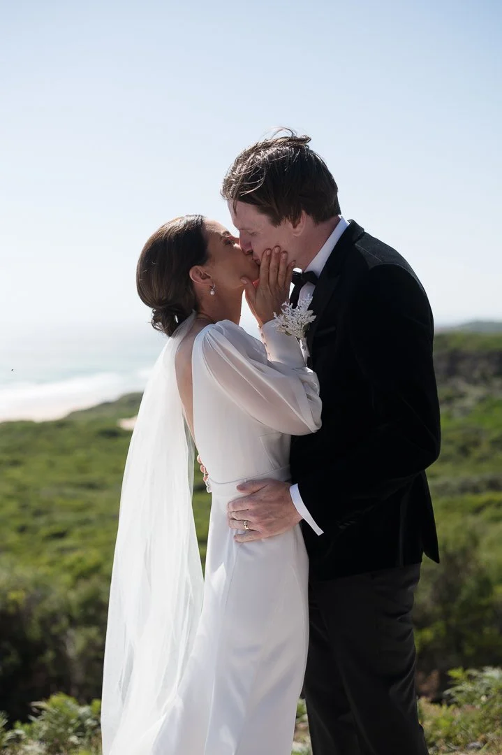 A bride and groom sharing a kiss outdoors near the coast with green landscape in the background.