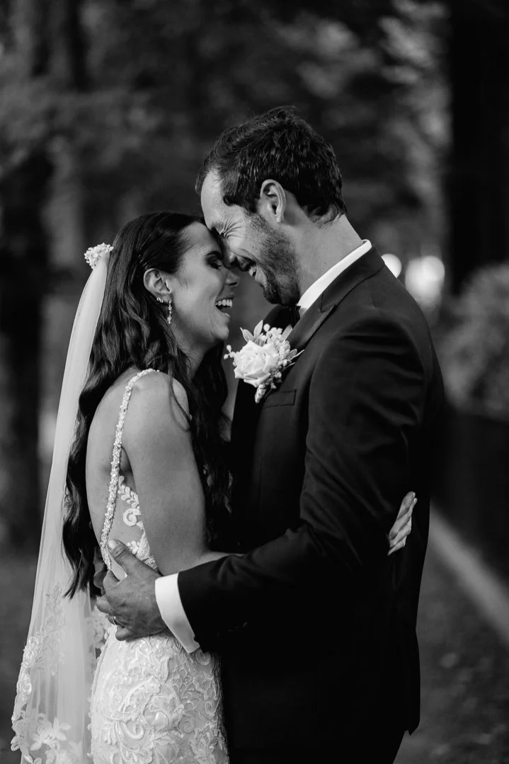 A black and white photo of a bride and groom embracing, smiling, and touching foreheads during their wedding, outdoors.