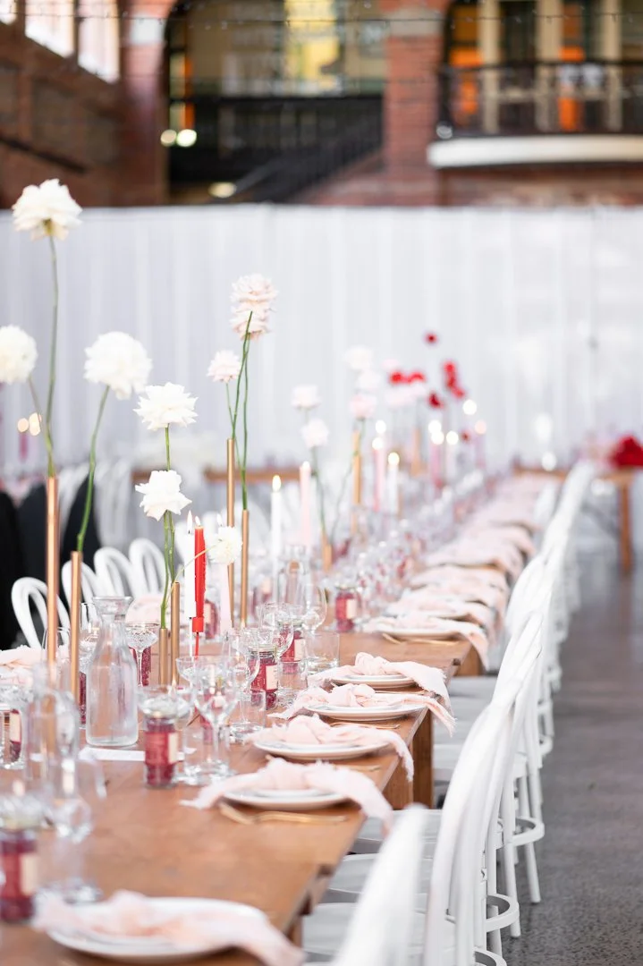 A long outdoor dining table set for a celebration with pink and white flowers, candles, glassware, and napkins, against a white fence and city buildings in the background.