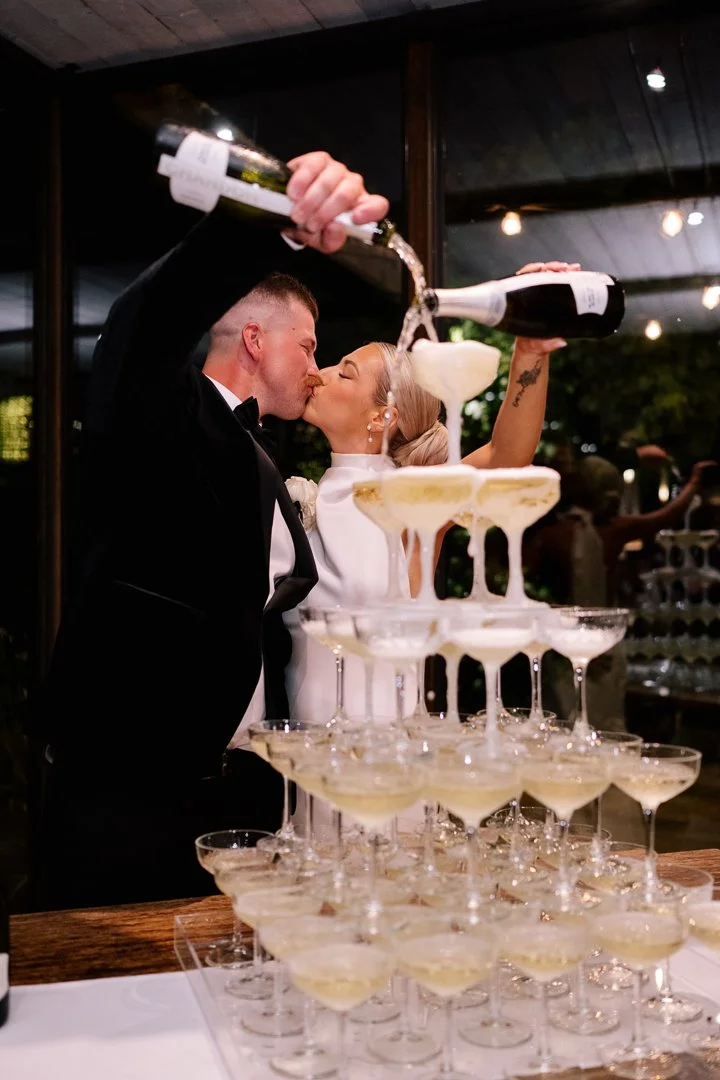 A bride and groom in wedding attire pouring champagne into a tower of glasses during their celebration.