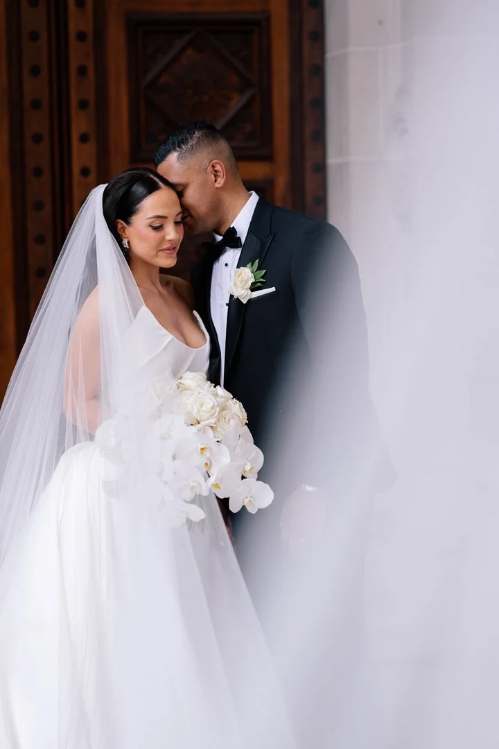 A bride and groom sharing a tender moment, with their foreheads touching. The bride is wearing a white wedding dress and veil, holding a bouquet of white flowers. The groom is dressed in a black tuxedo with a white shirt and a boutonniere. They are s