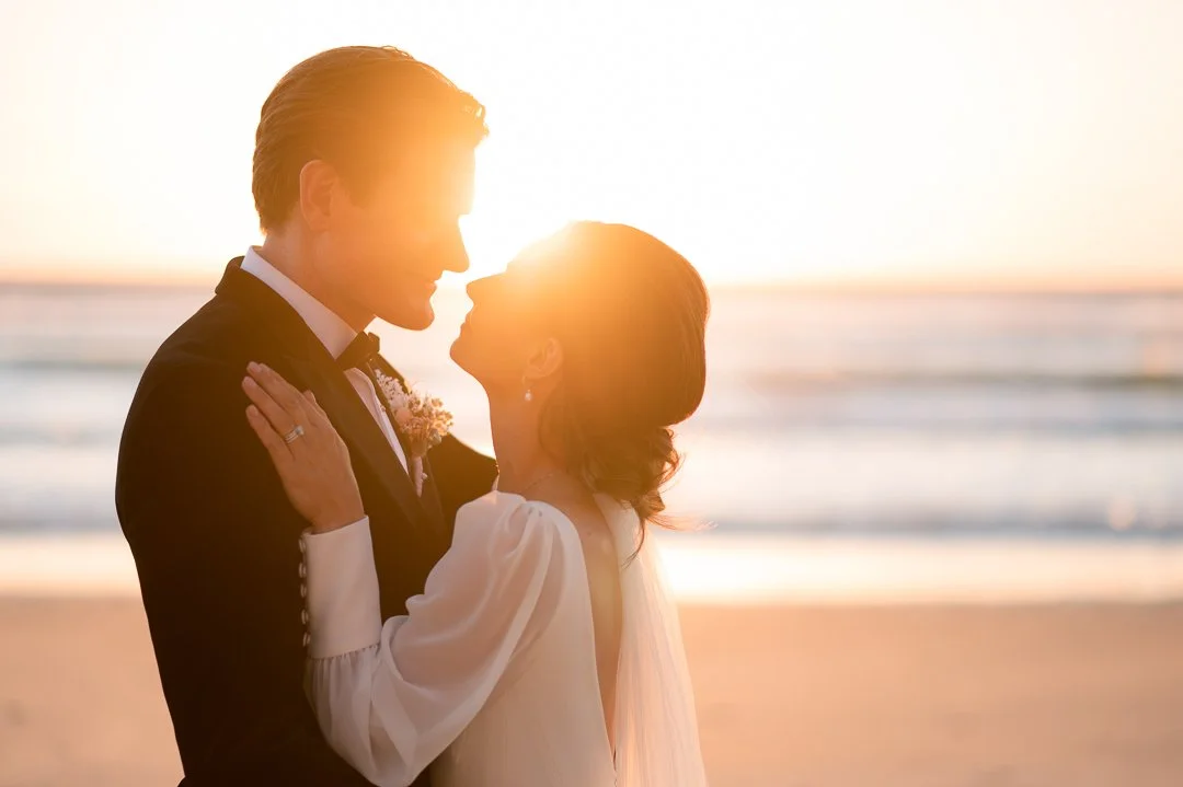 A couple in wedding attire sharing a kiss on the beach at sunset.
