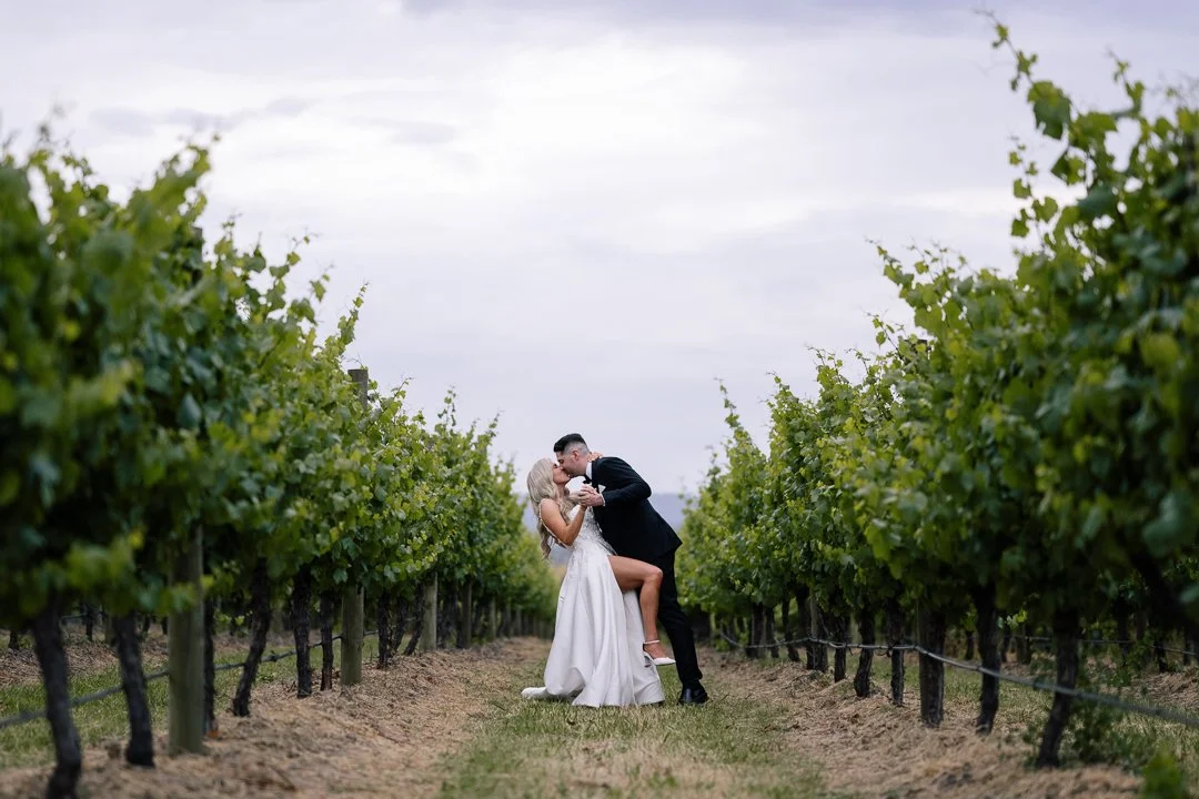 A newlywed couple sharing a kiss in a vineyard with lush green grapevines on either side.