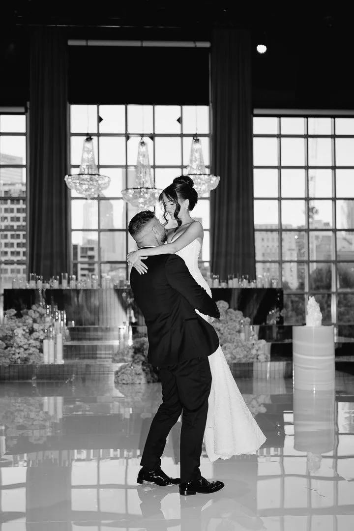 A black and white photo of a couple dancing at their wedding reception indoors, with large windows and chandeliers in the background.