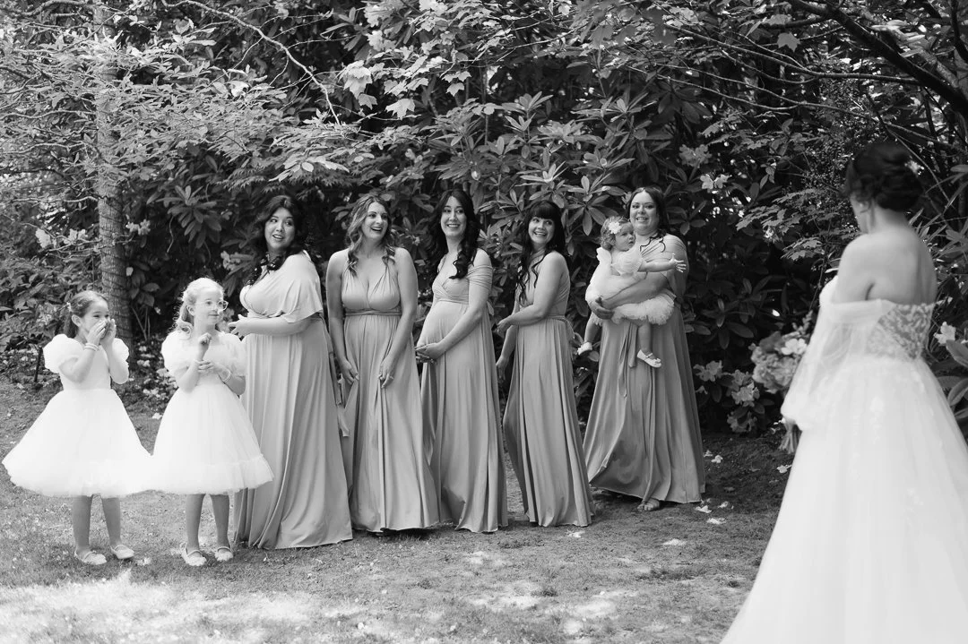 A group of women and young girls outdoors, with the women wearing matching long dresses and the girls in white dresses, all facing a woman in a strapless wedding gown, suggesting a wedding ceremony or preparation.