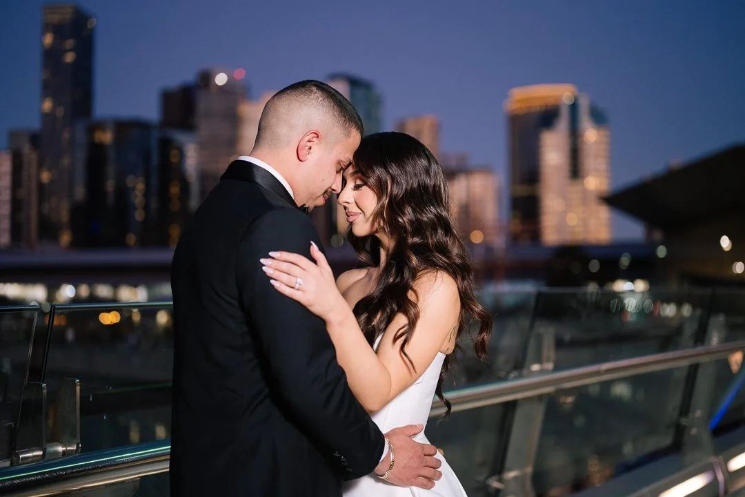 A couple dressed in formal attire, embracing closely on a rooftop at dusk with a city skyline in the background.
