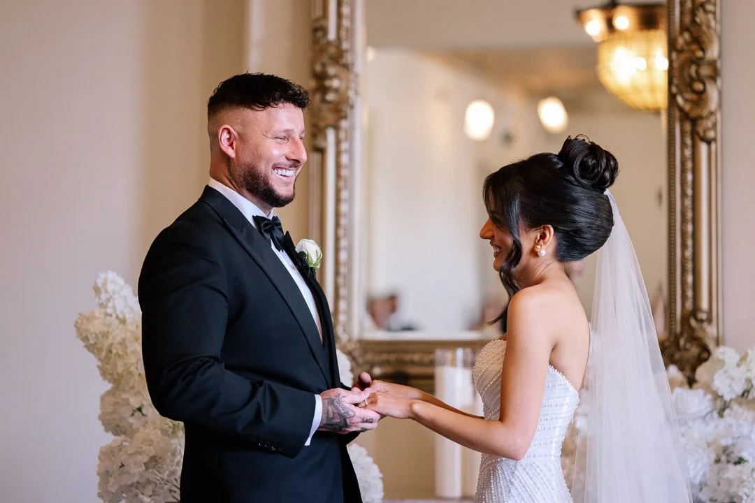 A bride and groom exchanging vows at their wedding ceremony, holding hands and smiling at each other in an elegant indoor setting.