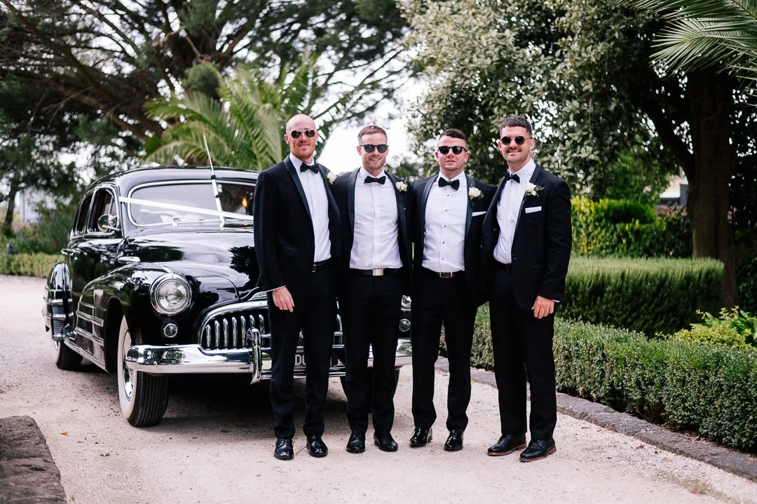 Four men dressed in tuxedos and sunglasses standing next to a vintage black car in a garden setting.